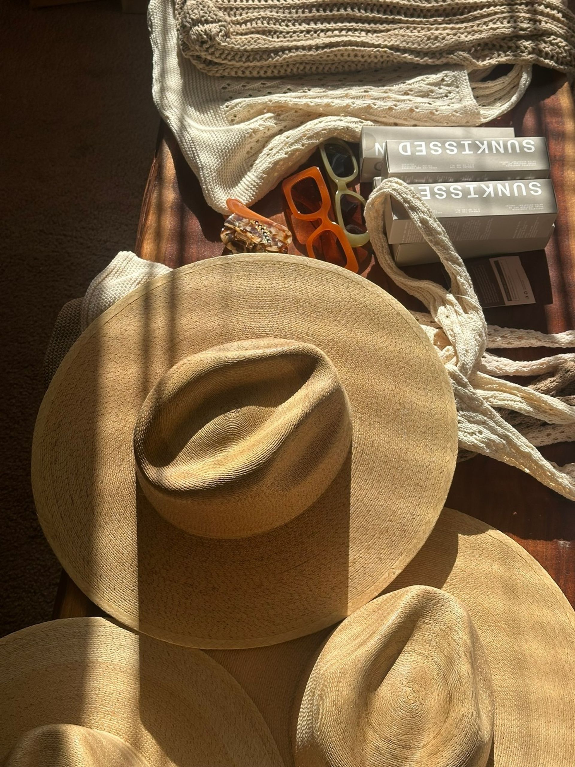 Sunlit flat lay of three wide-brim straw hats on a wooden table with cream knit scarves, two retro sunglasses, a tortoiseshell hair clip and stacked sunscreen boxes — summer beach accessories.