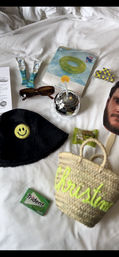 Pool-day flatlay on white hotel bed: straw tote with neon script, black bucket hat with yellow smiley patch, tortoiseshell sunglasses, packaged glitter swim ring, silver disco-ball cup with straw, travel-size tubes, Trident gum, small candy and hair clip.