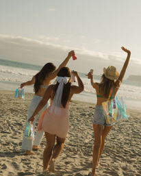 Three friends walking on a sunny sandy beach toward the ocean at golden hour, raising drinks and holographic tote bags — one wearing a bridal veil and bow for a bachelorette-style celebration.