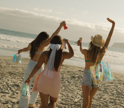 Three friends walking barefoot on a sandy ocean beach during golden hour, raising drinks and iridescent tote bags, one wearing a short veil — fun bachelorette-style summer celebration.