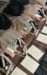 Beige mesh welcome bags on a rustic wooden table with black straw hats; each bag reveals sunscreen tubes, green-checkered cloth, boxed cosmetics, named hair clips and printed beach event itinerary cards — stylish beach party gift setup.