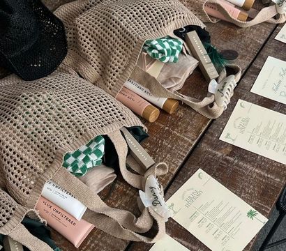Beige mesh welcome bags on a rustic wooden table with black straw hats; each bag reveals sunscreen tubes, green-checkered cloth, boxed cosmetics, named hair clips and printed beach event itinerary cards — stylish beach party gift setup.