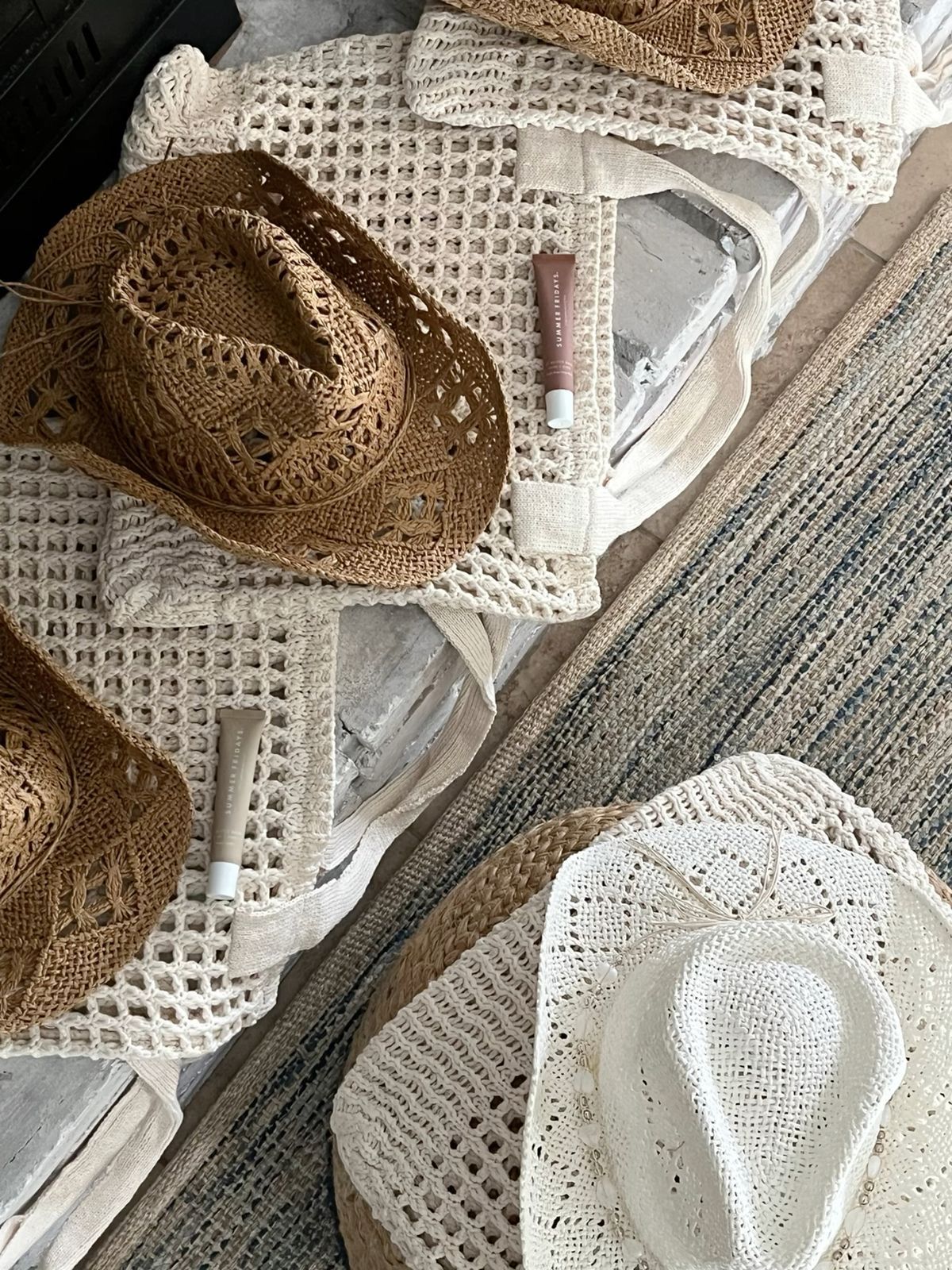 Flatlay of woven straw and white crochet sun hats, matching crochet tote bags and sunscreen tubes on a rustic coastal rug — boho beach essentials