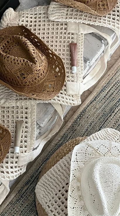 Flatlay of woven straw and white crochet sun hats, matching crochet tote bags and sunscreen tubes on a rustic coastal rug — boho beach essentials