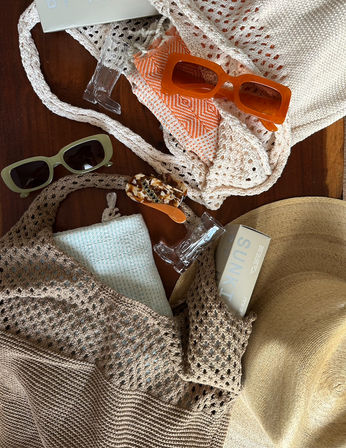 Summer beach flat-lay on wooden table: straw sun hat, beige crocheted totes, orange rectangular sunglasses, green shades, patterned pouch, mint towel, tortoiseshell hair clip and sunscreen box — casual beach-ready accessories.