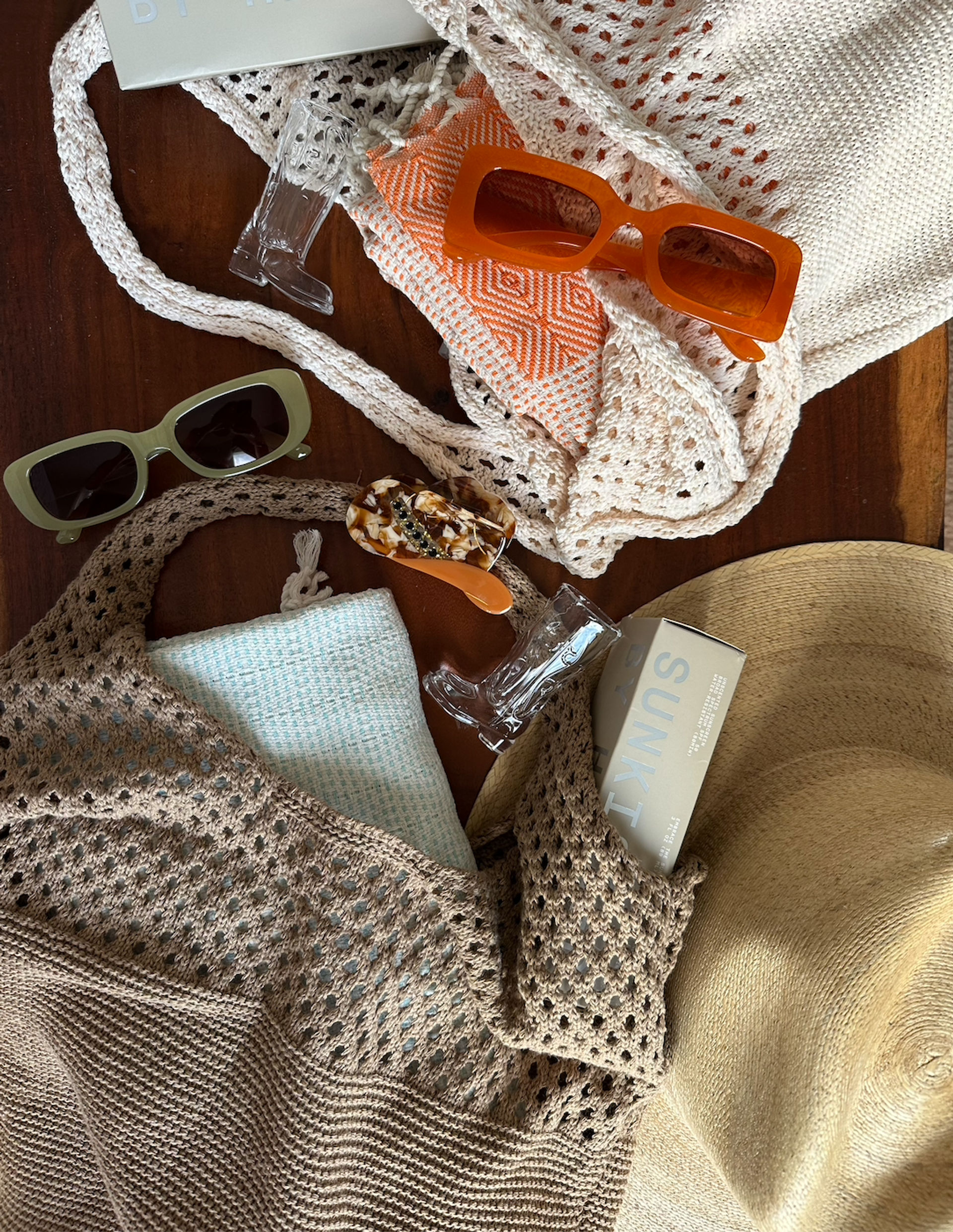 Summer beach flat-lay on wooden table: straw sun hat, beige crocheted totes, orange rectangular sunglasses, green shades, patterned pouch, mint towel, tortoiseshell hair clip and sunscreen box — casual beach-ready accessories.