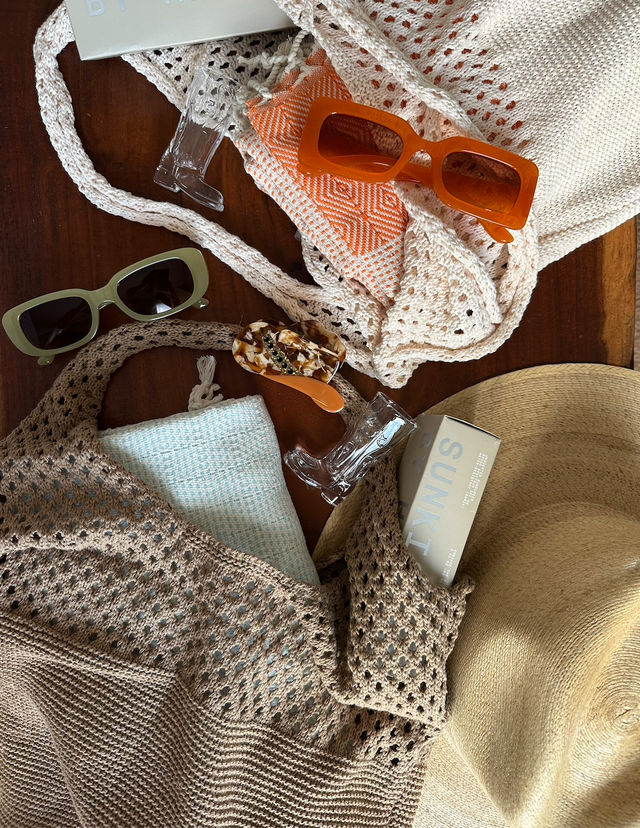 Summer beach flat-lay on wooden table: straw sun hat, beige crocheted totes, orange rectangular sunglasses, green shades, patterned pouch, mint towel, tortoiseshell hair clip and sunscreen box — casual beach-ready accessories.