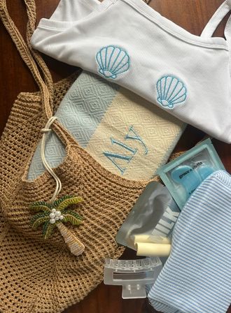 Flat-lay of summer beach essentials on wood: white ribbed bikini top with turquoise shell embroidery, monogrammed blue-and-beige towel, woven straw tote with beaded palm charm, striped pouch, clear hair claw and skincare sachets with under-eye gel patches.