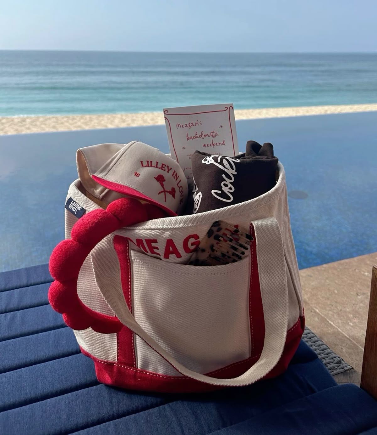 Red-and-white canvas beach tote holding a visor, rolled tee, hairbrush and a bachelorette-style invite, sitting poolside beside an infinity pool with a sandy beach and ocean horizon beyond.