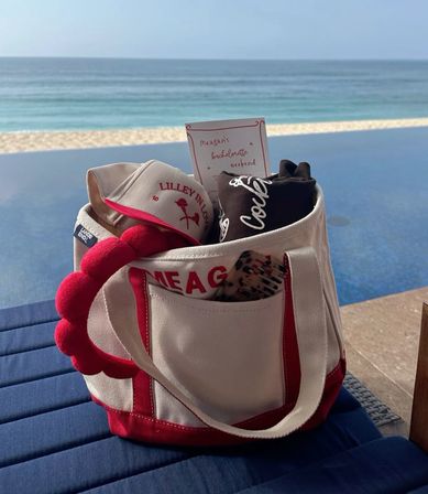 Red-and-white canvas beach tote holding a visor, rolled tee, hairbrush and a bachelorette-style invite, sitting poolside beside an infinity pool with a sandy beach and ocean horizon beyond.