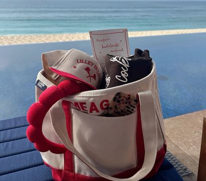 Red-and-white canvas beach tote holding a visor, rolled tee, hairbrush and a bachelorette-style invite, sitting poolside beside an infinity pool with a sandy beach and ocean horizon beyond.