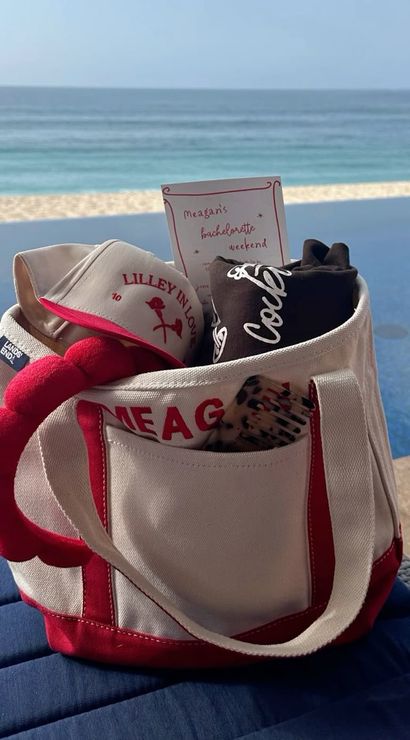 Red-and-white canvas beach tote holding a visor, rolled tee, hairbrush and a bachelorette-style invite, sitting poolside beside an infinity pool with a sandy beach and ocean horizon beyond.