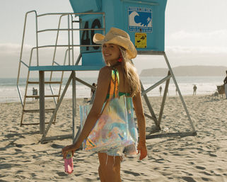 Smiling woman in a straw cowboy hat and denim shorts carrying a holographic tote and drink past a blue lifeguard tower on a sunny sandy beach with ocean waves.