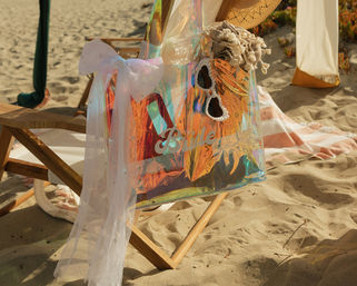 Iridescent tote bag labeled “Bride” with pearl-trim heart sunglasses, straw hat and sheer white bow draped over a wooden beach chair on a sunlit sandy beach
