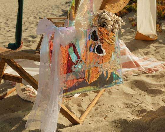Shimmering iridescent bridal tote labeled 'Bride' with pearl‑trim heart sunglasses and tulle bow draped over a wooden beach chair on a sunlit sandy shore, beach wedding vibes.
