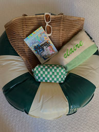 Beach-ready flat lay: brown mesh tote with white sunglasses, holographic hangover kit, green checked toiletry pouch and embroidered towel on a green-and-cream inflatable pool float.