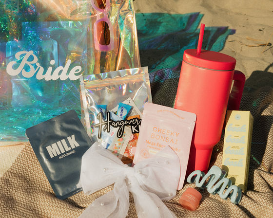 Sunny bachelorette beach flatlay with iridescent tote labeled "Bride", pink sunglasses, white bow, moisturizing face mask packet, hangover kit pouch, energizing patch sachet, coral travel tumbler with straw, sunscreen box, lip balm and blue hair claw on a patterned blanket