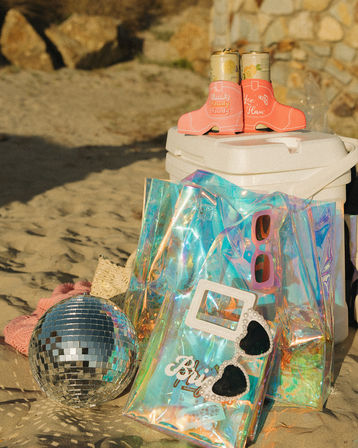 Sunny beach still-life: white cooler topped with two canned drinks in pink cowboy-boot can koozies, iridescent holographic tote holding pink sunglasses and pearl heart shades, small disco ball and knit hat on sand.