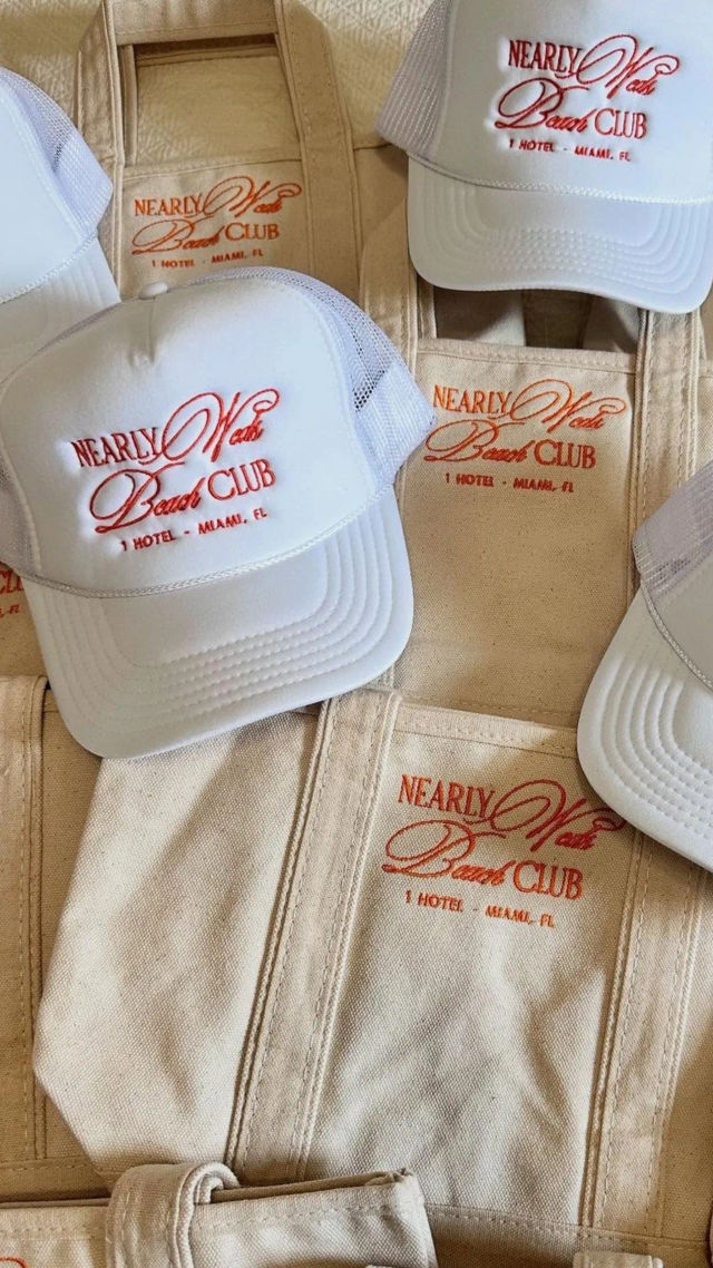 White mesh trucker hats and beige canvas tote bags embroidered with red beach-club style logo and Miami, FL text, arranged flat for merch display