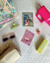 Vacation-ready flat lay on white quilted bedspread with a holographic tote, lime green spiral hair clip, resealable hangover kit pouch, neon pink insulated travel tumbler, gray rectangular sunglasses, folded beige woven throw, pink energy supplement sachet, yellow product box and small lip balm.