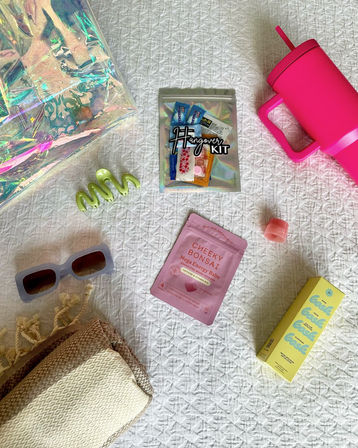 Beach day essentials flat lay on white quilt: iridescent tote, holographic hangover kit pouch, hot pink insulated tumbler, retro sunglasses, green hair claw, pink energy sachet, lip balm, yellow sunscreen box and folded woven beach towel.