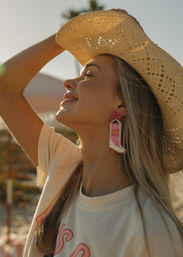 Sunlit profile of a smiling woman at the beach wearing a woven straw hat and colorful beaded cowboy-boot earring, coastal golden-hour summer vibe.