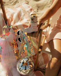 Sunlit beach bachelorette setup: woman in a pink dress holding a canned drink in a pink cowboy-boot koozie reading 'gettin' hitched', iridescent bride gift bag, pearl sunglasses, disco ball on a striped towel.