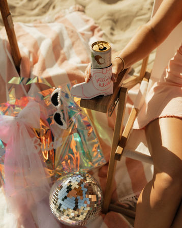Sunny beach bachelorette scene: woman in a pink dress seated on a wooden chair holding a canned drink in a white cowboy-boot can cooler that reads “Gettin’ hitched,” with a striped beach towel, iridescent gift bag, pearl sunglasses, tulle veil and disco ball on the sand.