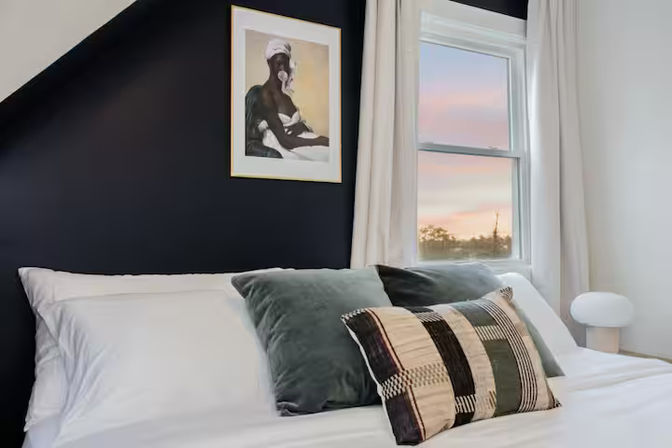 Cozy modern bedroom with white linens, velvet green pillows and a patterned throw pillow on a bed against a navy accent wall, framed artwork above, a window with a pink sunset view and a small round bedside lamp.
