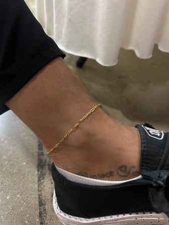 Close-up of an ankle wearing a delicate gold anklet with a faint script tattoo, black slip-on sneaker and white sock on a concrete floor beneath a white tablecloth.