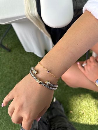 Close-up of a wrist with a twisted silver-and-gold cuff and delicate gold chain bracelet with a small purple stone, light pink nails, on an outdoor artificial-grass patio near a white table.