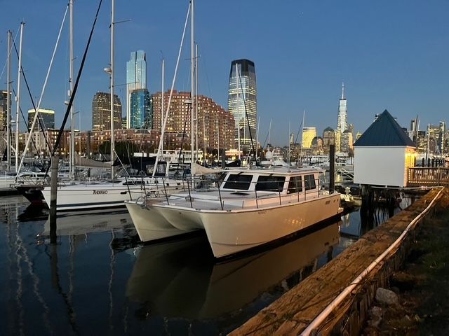 New York City marina at dusk with a docked white motorboat and sailboats, calm water reflecting illuminated skyscrapers of the skyline.