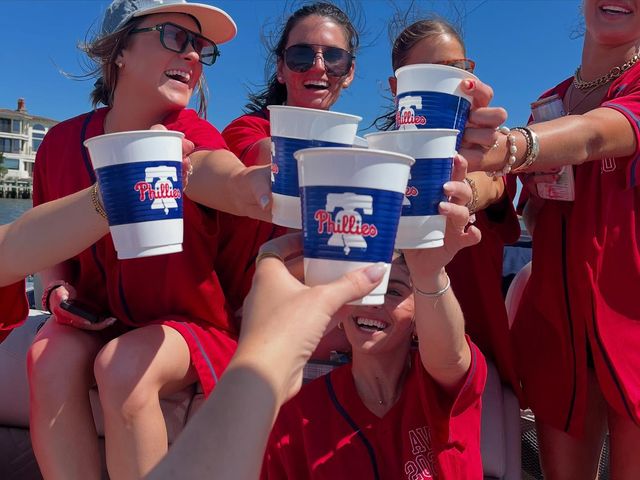 Group of friends in red jerseys cheering and clinking Phillies-branded cups on a sunny boat party by the water