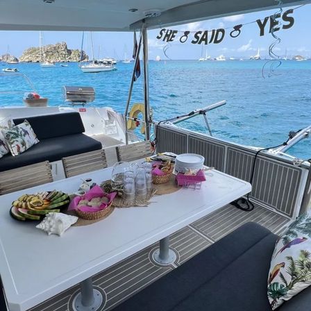 Yacht deck setup for an engagement celebration: table with plates, glasses, tropical fruit and snacks, cushioned seating and a ‘She Said Yes’ banner, turquoise bay with anchored sailboats and rocky islets in the background.