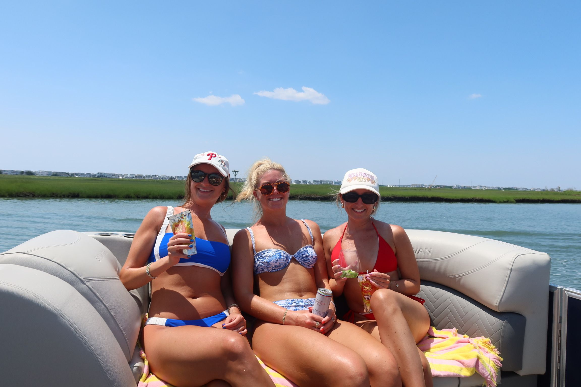 Three friends in colorful bikinis and caps enjoying drinks on a pontoon boat near a coastal marsh under a bright summer sky