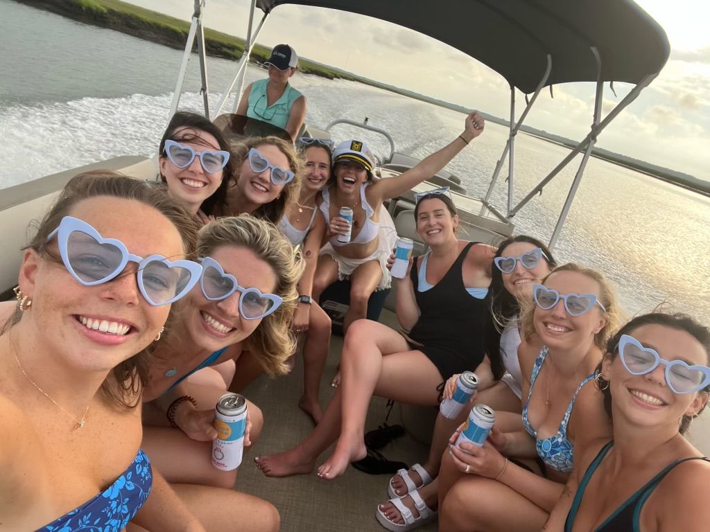 Smiling group of women wearing matching light-blue heart-shaped sunglasses on a pontoon boat, holding canned drinks during a sunlit coastal inlet cruise