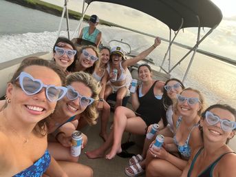 Smiling group of women wearing matching light-blue heart-shaped sunglasses on a pontoon boat, holding canned drinks during a sunlit coastal inlet cruise