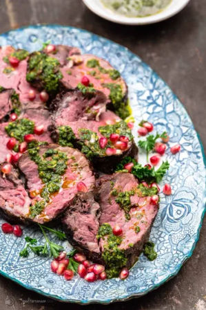 Sliced medium-rare beef tenderloin drizzled with vibrant green herb sauce, garnished with pomegranate arils and parsley on a blue patterned serving plate