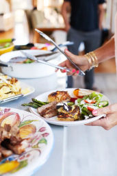 Hand with tongs serving a colorful buffet plate of grilled asparagus, sliced roast, roasted potatoes and fresh mixed salad at a casual indoor gathering.