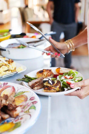 Hand with tongs serving a colorful buffet plate of grilled asparagus, sliced roast, roasted potatoes and fresh mixed salad at a casual indoor gathering.