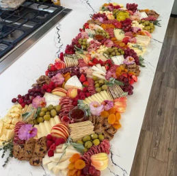 Long colorful charcuterie grazing board on a white marble kitchen island with assorted cheeses, cured meats, crackers, olives, grapes, sliced apples, dried apricots and edible flowers for a party spread