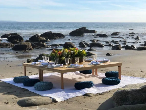 Low wooden table set for a beach picnic on a sandy shore with blue round cushions on a white rug, glassware and a vibrant flower centerpiece against a rocky ocean horizon