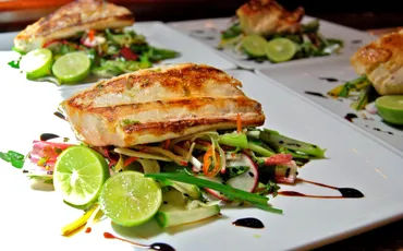 Grilled fish fillet perched on a colorful julienned vegetable salad, garnished with halved limes and a balsamic drizzle on white restaurant plates.