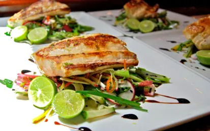 Grilled fish fillet perched on a colorful julienned vegetable salad, garnished with halved limes and a balsamic drizzle on white restaurant plates.