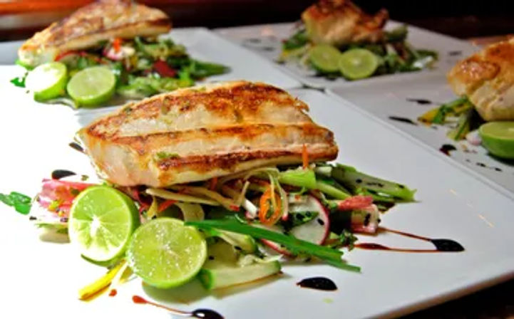 Grilled fish fillet perched on a colorful julienned vegetable salad, garnished with halved limes and a balsamic drizzle on white restaurant plates.