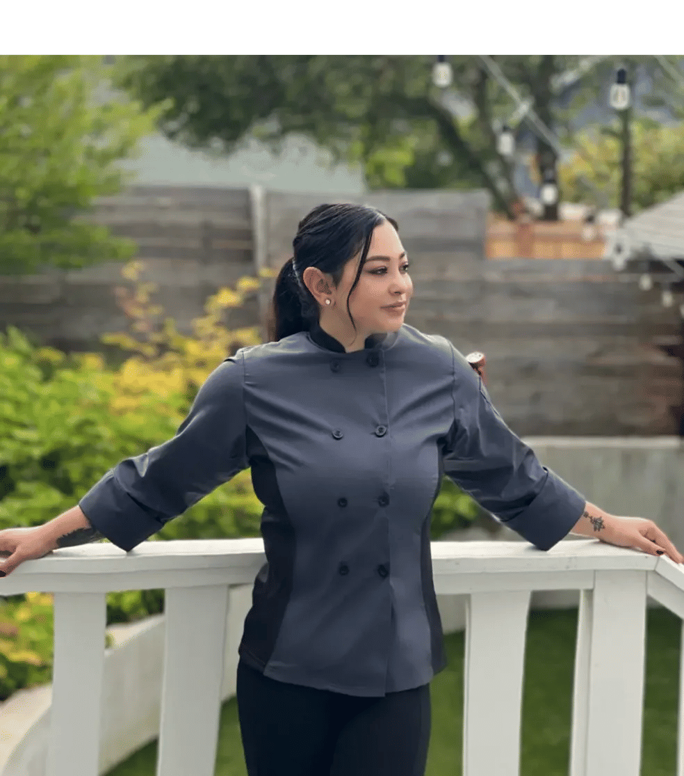 Person in a gray chef jacket leans on a white deck railing, hair tied back, looking to the side with a backyard garden and string lights in the background.