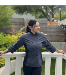 Person in a gray chef jacket leans on a white deck railing, hair tied back, looking to the side with a backyard garden and string lights in the background.