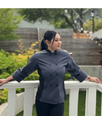 Person in a gray chef jacket leans on a white deck railing, hair tied back, looking to the side with a backyard garden and string lights in the background.