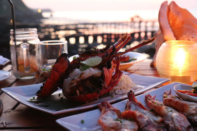 Candlelit beachside seafood spread with grilled lobster tails and prawns on white plates, mason jar drink and a pier silhouetted against a pink ocean sunset