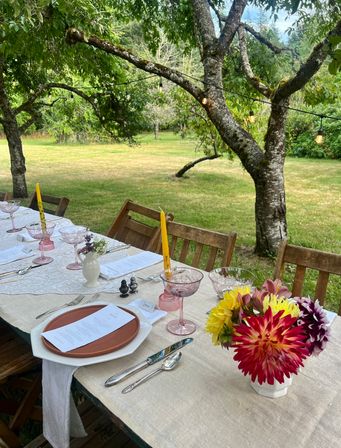 Outdoor garden dining table in a grassy backyard under leafy trees, rustic wooden chairs, linen tablecloth, pink stemmed glasses, yellow taper candles, menus on terracotta plates, fresh dahlia bouquet and string lights overhead